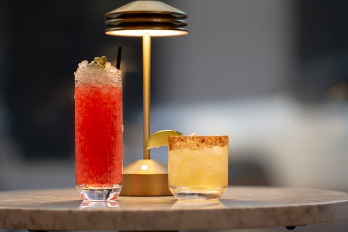 A tall glass filled with ice and red liquid sits on a table next to a shorter glass with a lime wedge and yellow liquid. A lamp glows behind them.