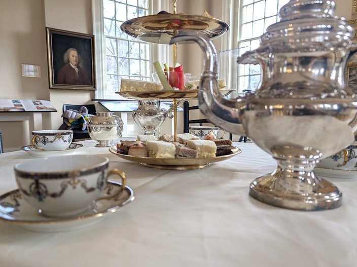 A white, gold and blue teacup and saucer (left) sits on a table with a silver teapot (right) and a three-tiered tray of sandwiches.