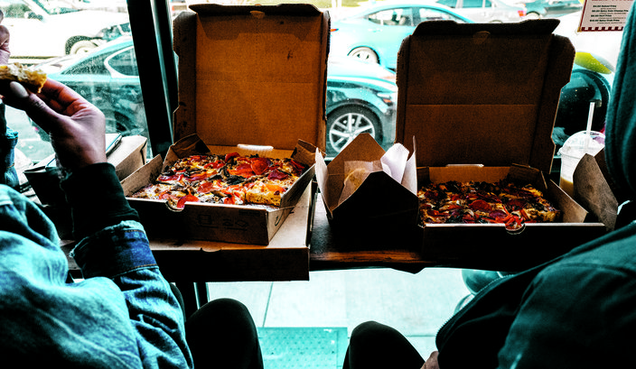 Over the shoulder shot of two people eating two pizzas in cardboard boxes.