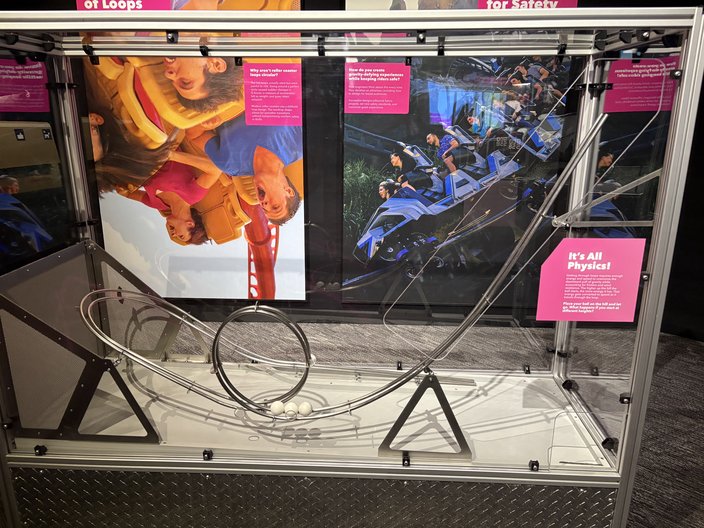 Roller coaster loop model in a glass case with pink signage and photos behind it