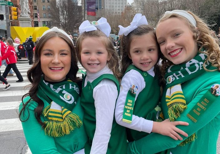 Four Irish step dancers in green and white costumes in front of a Philadelphia street.