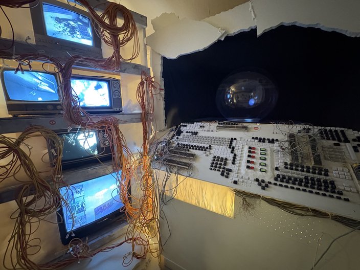 Five TV monitors stacked on shelves surrounded by wires; a damaged keyboard is to the right