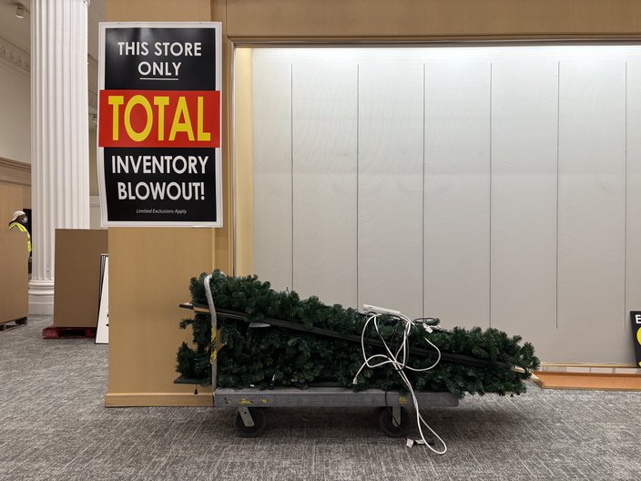 A fake Christmas tree lays tied to a metal cart under a sign reading 'THIS STORE ONLY TOTAL INVENTORY BLOWOUT!'