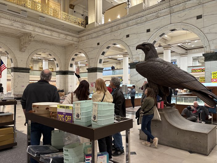 Shoppers on a department store floor with a table of baskets for sale and a large eagle statue