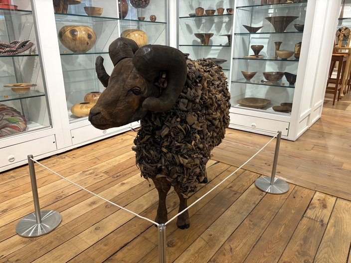 A wooden sculpture of a ram inside a museum with glass and white display cases behind it