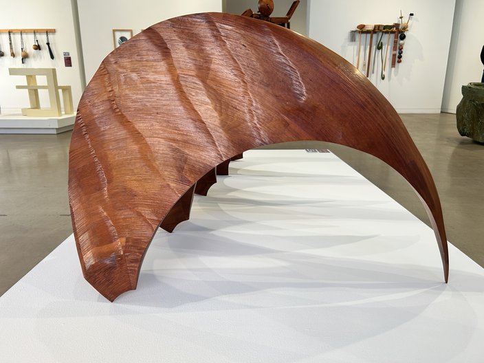 A curved wooden sculpture with textured waves sits atop a white floor