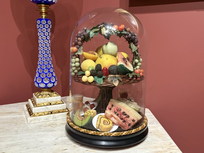 A glass case of wax fruit sits on a table, with a blue lamp base behind it