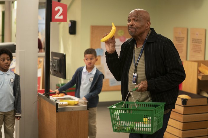 Man in a black jacket and blue lanyard holds up a banana in a play grocery store. He holds a green shopping basket in his other hand.