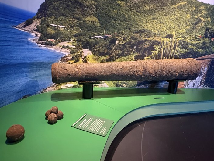 An old cannon is displayed on a green ledge with a photo of the Caribbean island of Sint Eustatius behind it