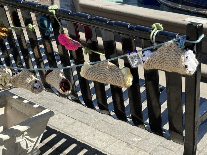 Knitted cannoli hang from a string across an iron bench.