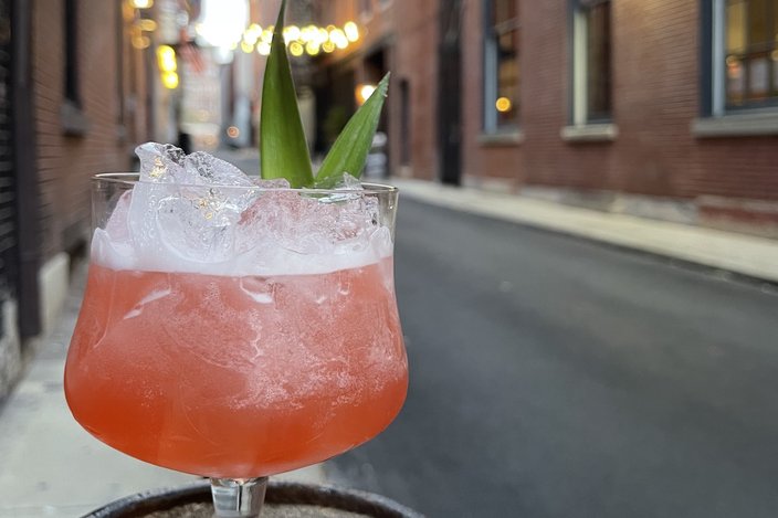 Pink cocktail in a class with ice and green garnish. A city street is visible in the background.