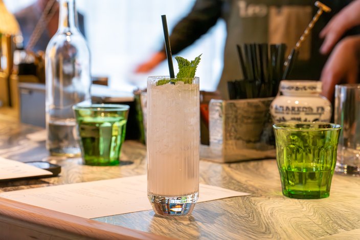Pale cocktail in a tall glass with ice, mint leaves and a black straw on a tabletop.