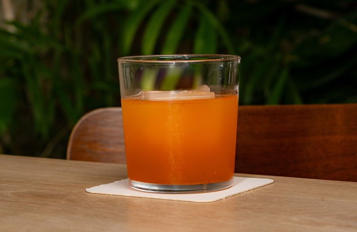 An orange-colored drink in a rocks glass with a white napkin underneath. It sits on a tabletop with a green plant in the background.