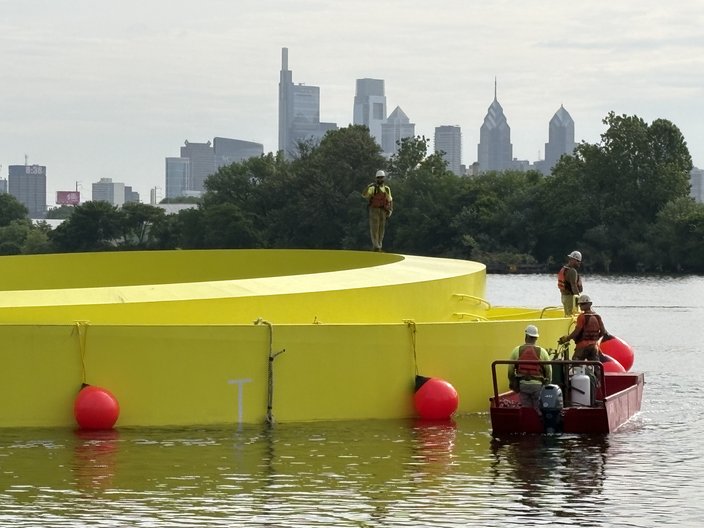 Workers on the Schuylkill River inspect a bright yellow floating observation deck. The Philadelphia skyline is in the background