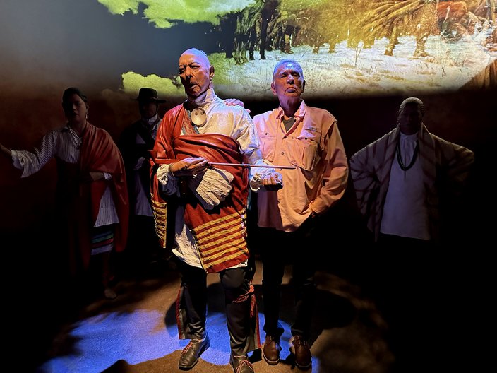 A man in a salmon-colored shirt (right) stands next to a painted statue of a Native American man (left) on a stage.