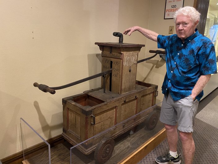 A man in a blue button-down shirt and shorts gestures to a wooden 1792 hand engine used to fight fires.