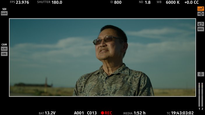 A man in sunglasses and a gray shirt stands in front of a blue sky with light clouding.