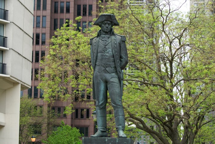 A bronze statue of a man in 18th century dress, with a tree and buildings in the background.