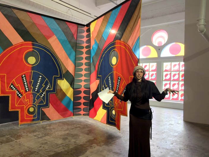 A woman in black stands in front of a large, multi-colored piece of fabric hanging from a ceiling