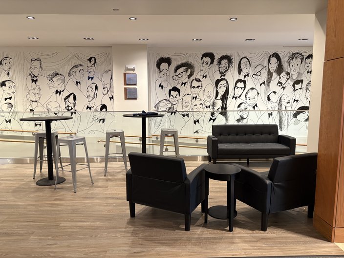 A movie theater lobby space with black chairs, tables and a couch. The wall behind is painted with illustrations by Al Hirschfeld.