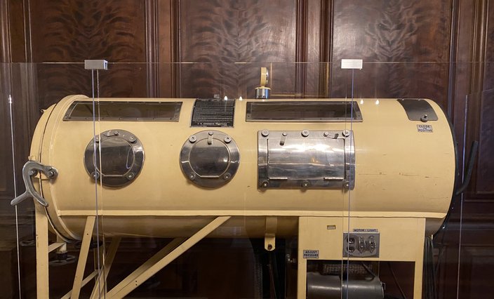 A tan Emerson iron lung in a glass display case in front of dark wood paneling