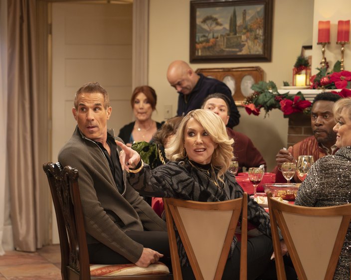 A blond woman seated at a table points her finger. Four other people sit alongside her, with another couple in the background.