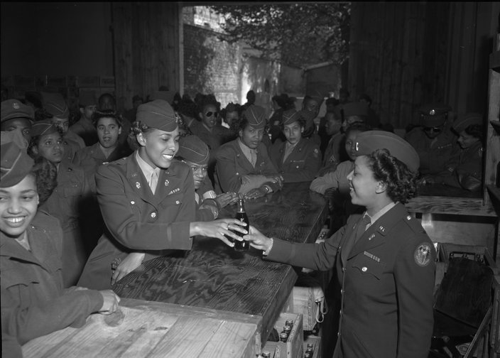 A black-and-white photo of Black women in military uniforms gathered around a bar. One passes a glass bottle of Coke to another in the foreground.