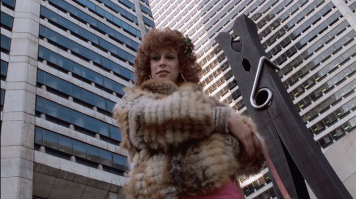 A woman with red hair wearing a fur coat stands in front of the 'Clothespin' statue in Philadelphia.