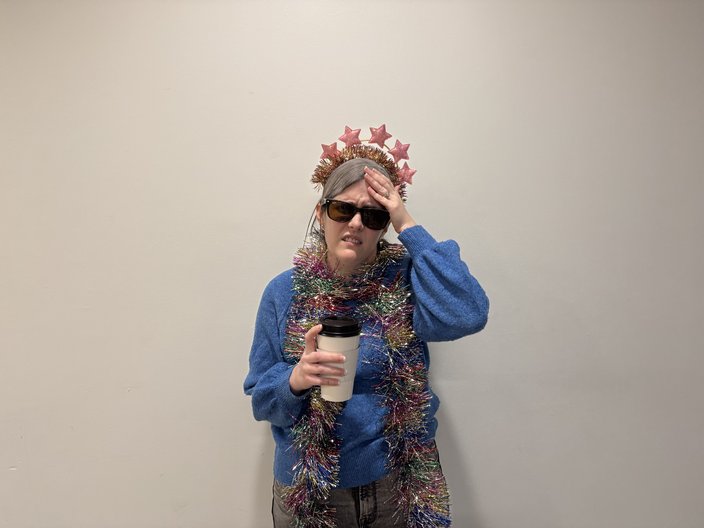 Woman in blue sweater, sunglasses and party headband holds a coffee cup in one hand and clutches her forehead with the other