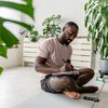 man sitting on a yoga mat at home, writing in a notebook after a workout