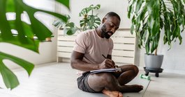 man sitting on a yoga mat at home, writing in a notebook after a workout