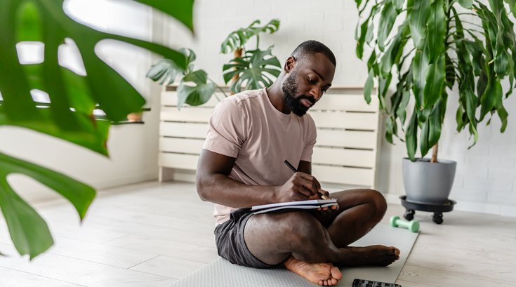 man sitting on a yoga mat at home, writing in a notebook after a workout