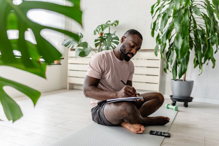 man sitting on a yoga mat at home, writing in a notebook after a workout