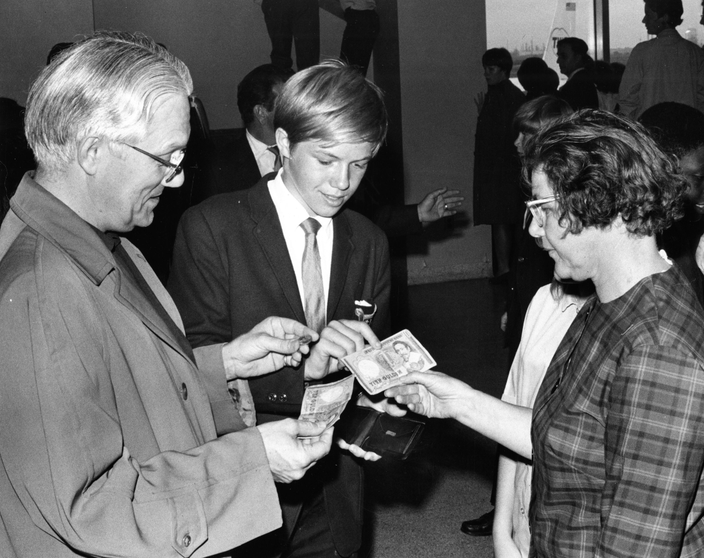 Black-and-white photo of a boy in a suit (center) showing money to a man and a woman in glasses