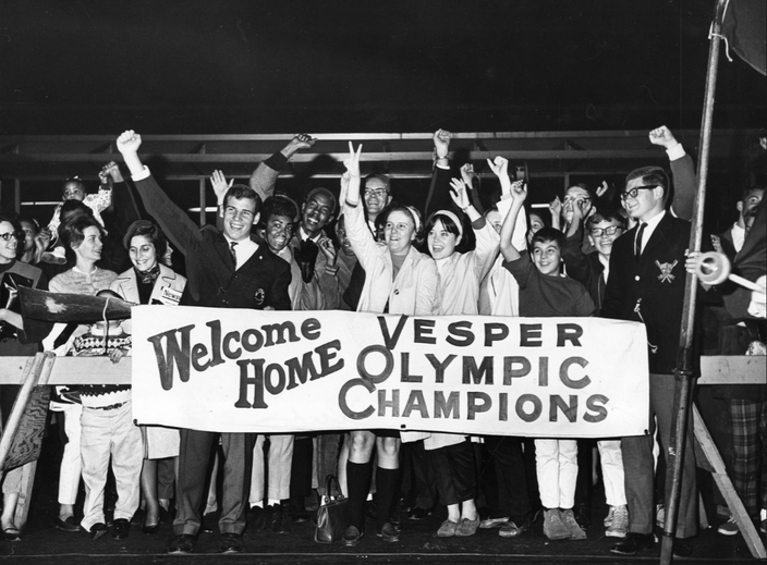 Black-and-white photo of fans cheering the Vesper Boating Club at the Philly airport. They hold a sign reading 'Welcome Home Vesper Olympic Champions'